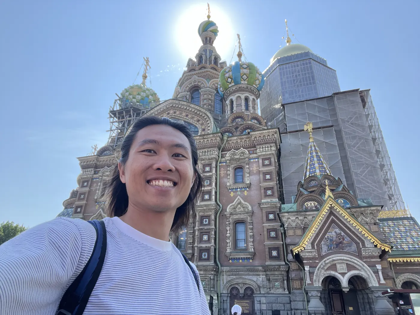 Shin selfie in front of the Church of the Saviour on Spilled Blood with its colourful onion domes under blue summer sky, partially covered in restoration scaffolding
