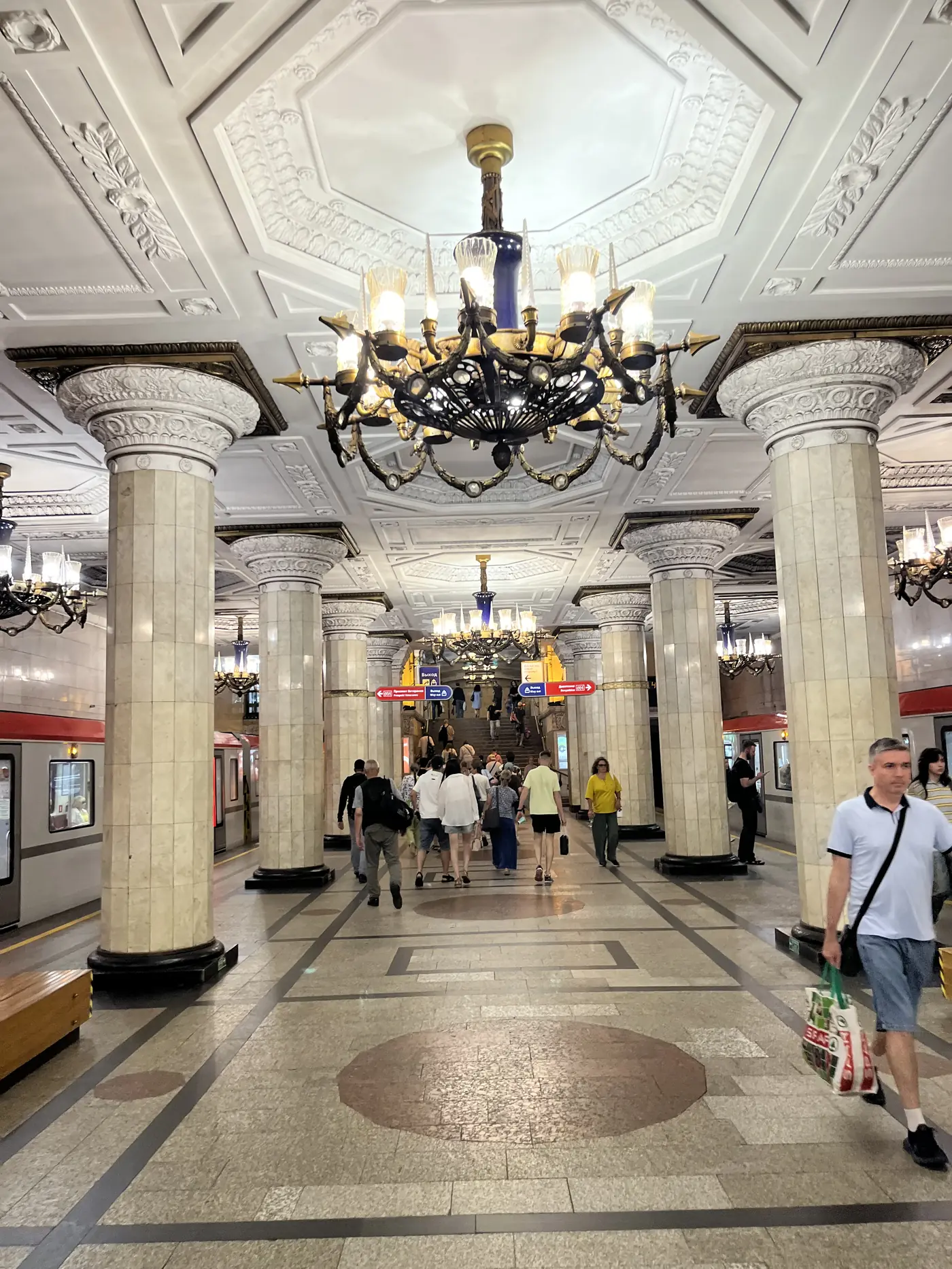 Grand St. Petersburg metro station with marble columns, ornate ceiling medallions, chandeliers, and commuters walking beneath