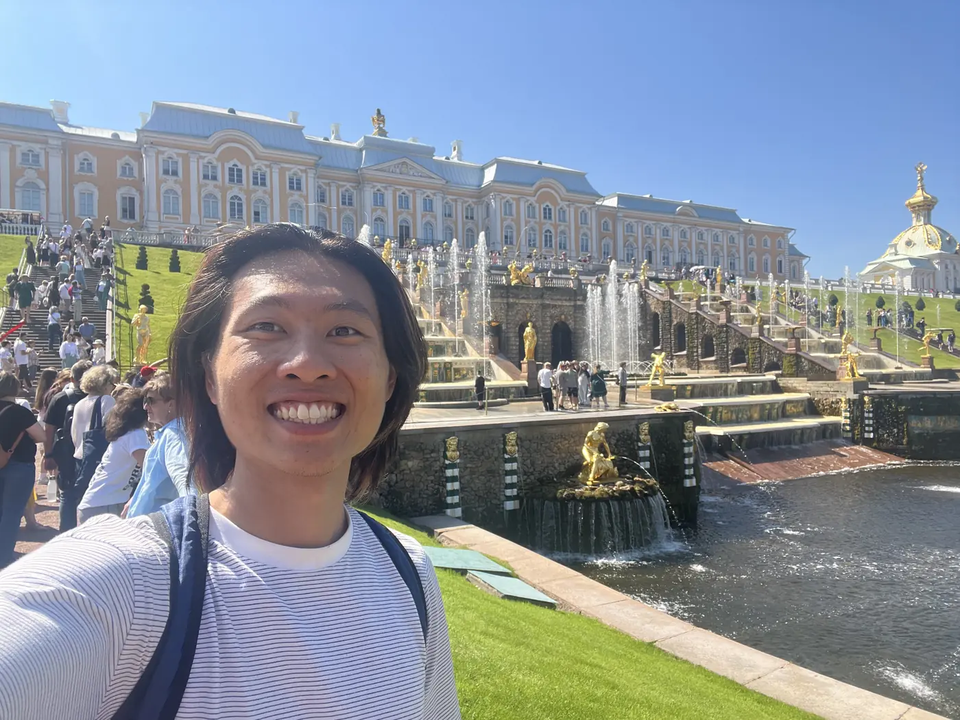 Shin selfie at Peterhof Palace with the Grand Cascade's golden statues and fountains cascading down behind him toward the Gulf of Finland