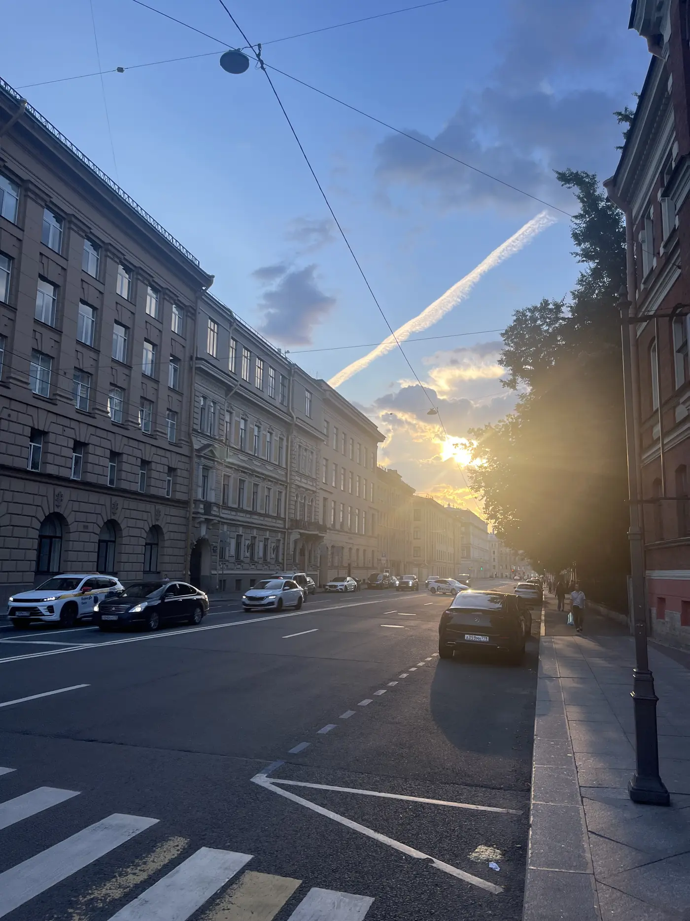 St. Petersburg street at golden hour, sun flaring between apartment buildings and trees, cars parked along the curb