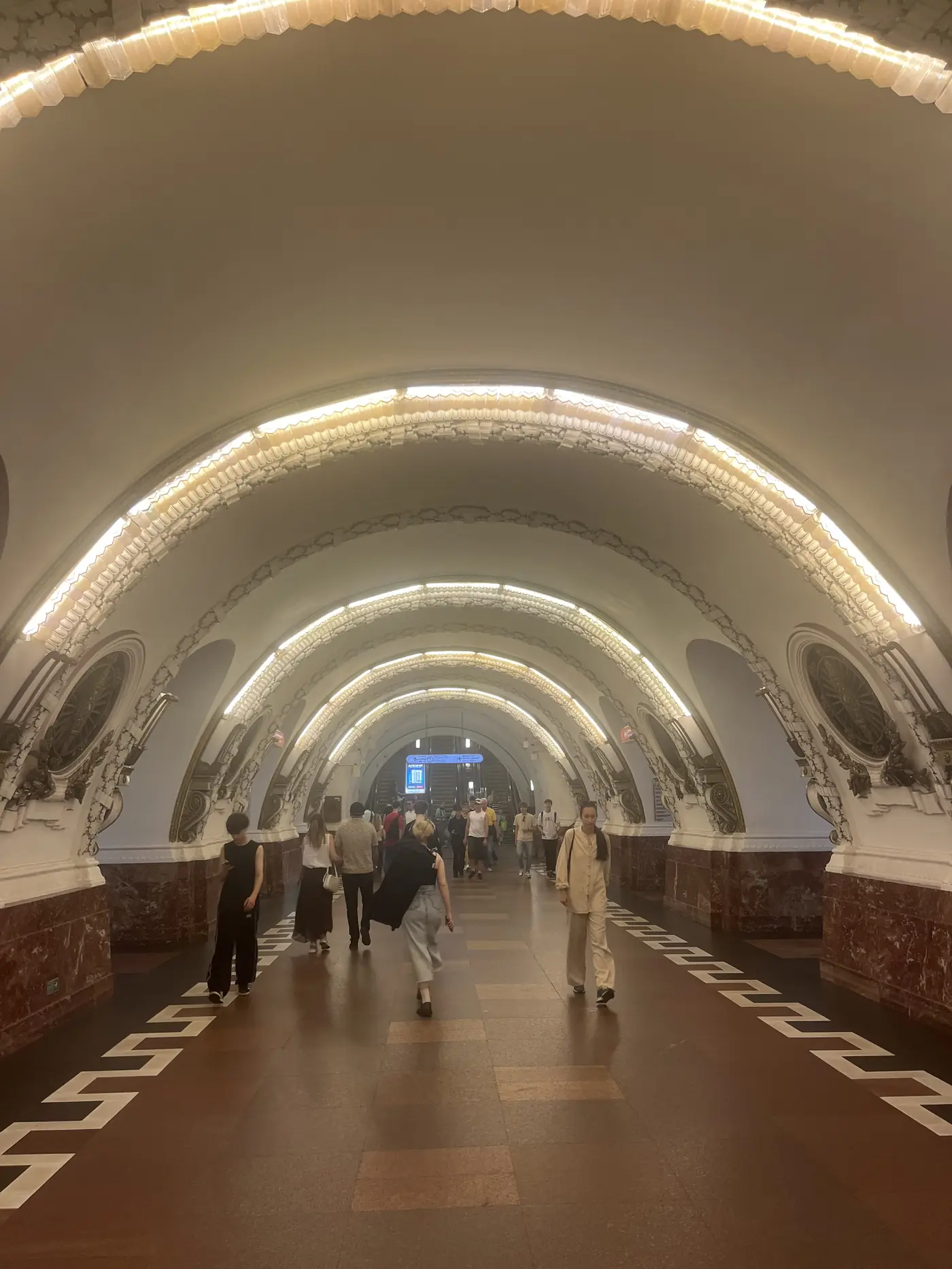 Ornate St. Petersburg metro station with a barrel-vaulted white ceiling lit by rings of warm light, red marble columns and decorative plaster medallions, commuters walking through