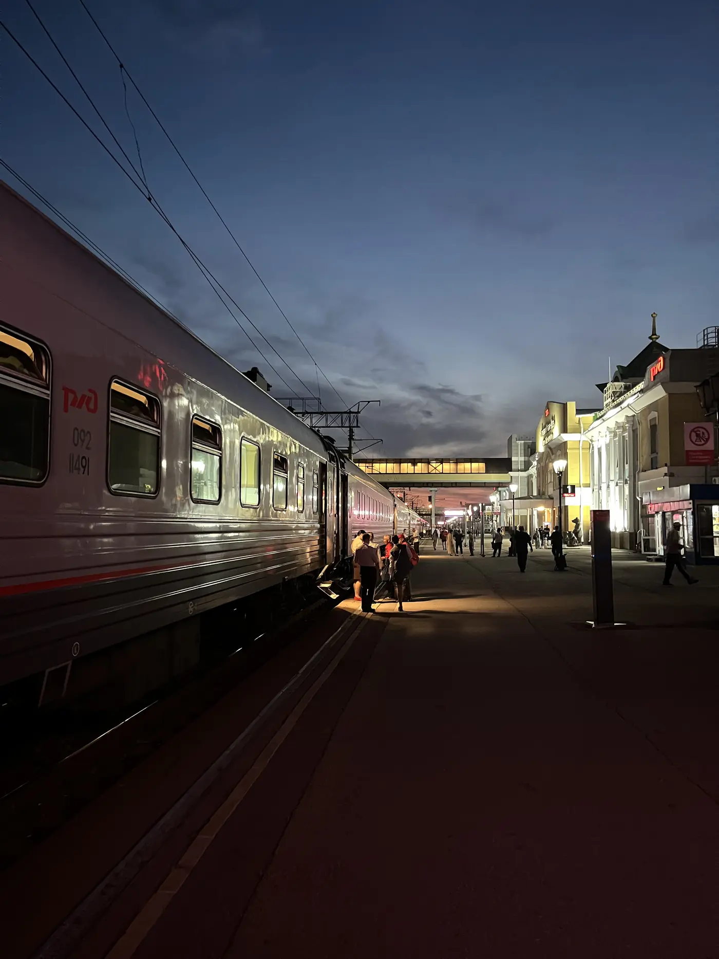 Trans-Siberian Railway silver RZhD train at Ulan-Ude platform at night, warm station lights, passengers on the platform