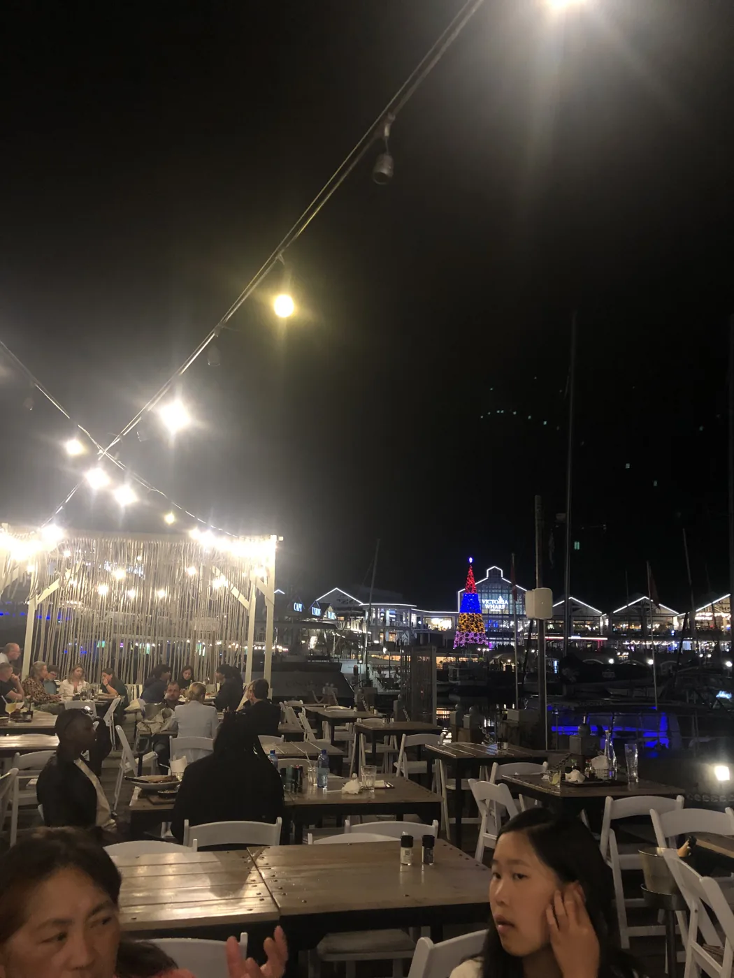 Outdoor waterfront restaurant at night with V&A Waterfront lit up and a Christmas tree glowing behind