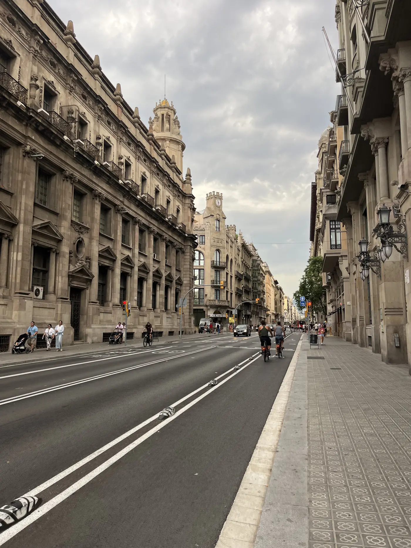 Wide Barcelona boulevard lined with neoclassical stone buildings, cyclists on the bike lane and a clock tower in the distance