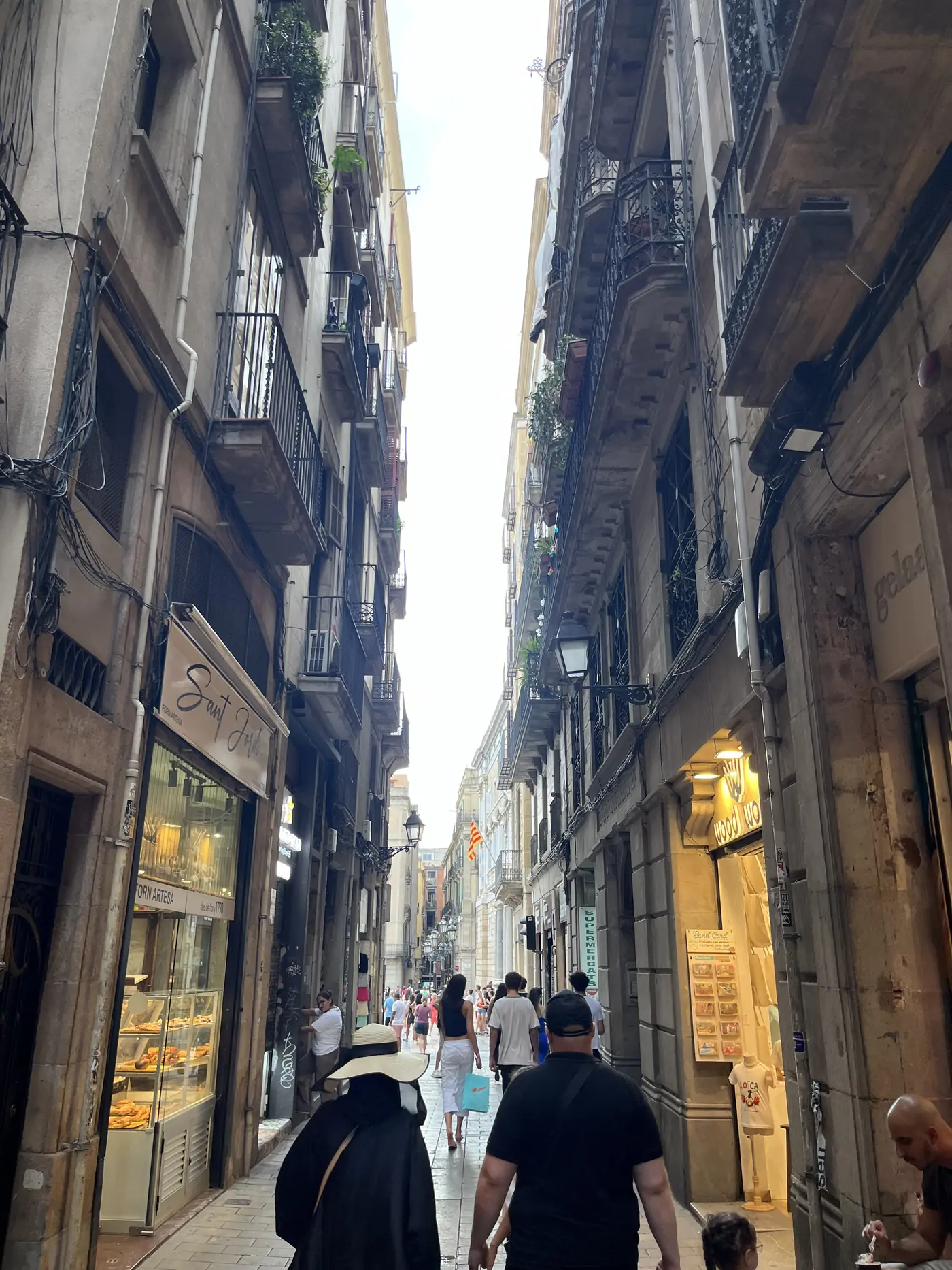 Narrow medieval alley in Barcelona's Gothic Quarter lined with tall stone buildings, balconies, and small shops, crowds walking through