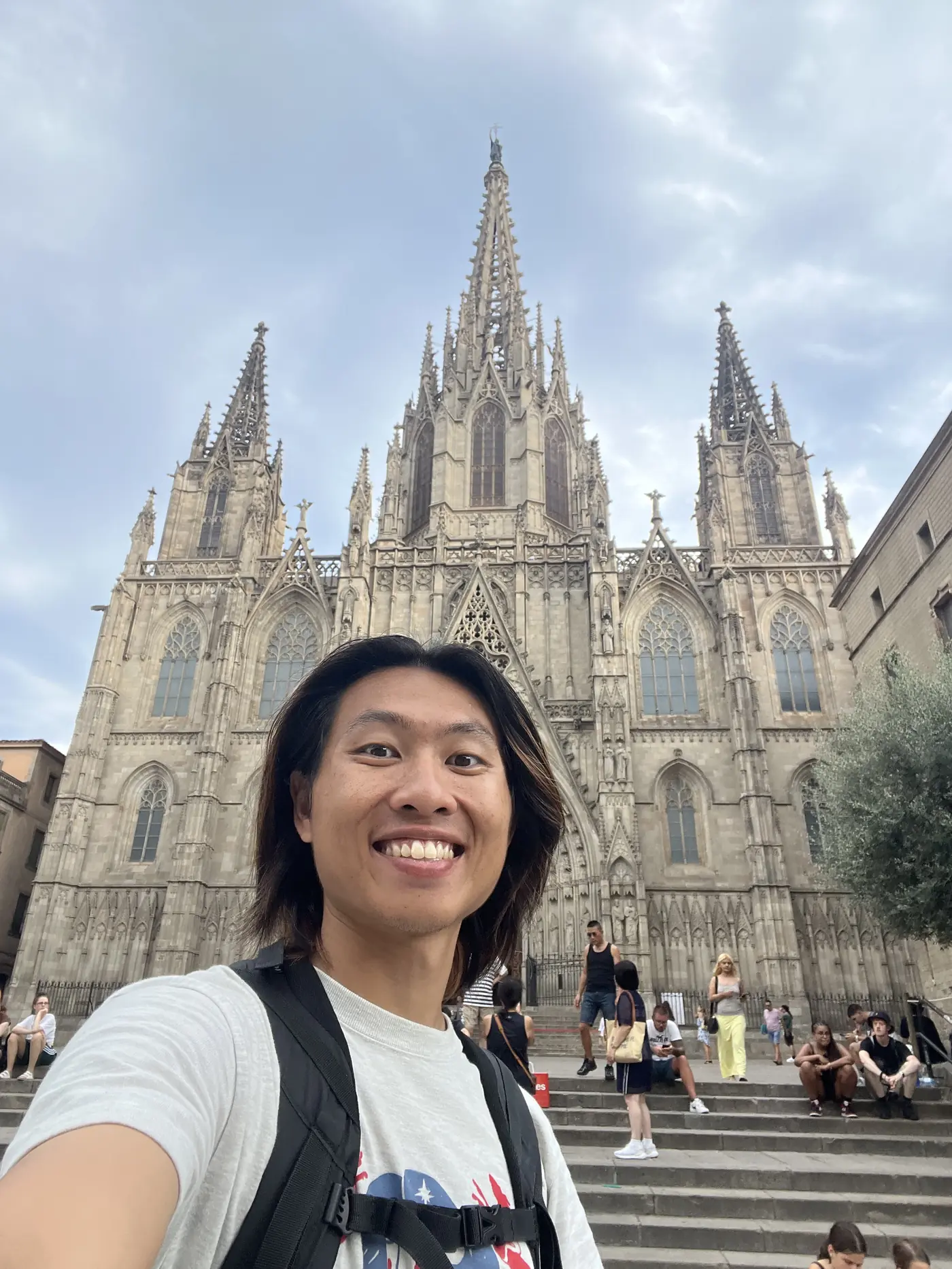 Shin selfie in front of Barcelona Cathedral's Gothic spire and facade, with people sitting on the steps behind him
