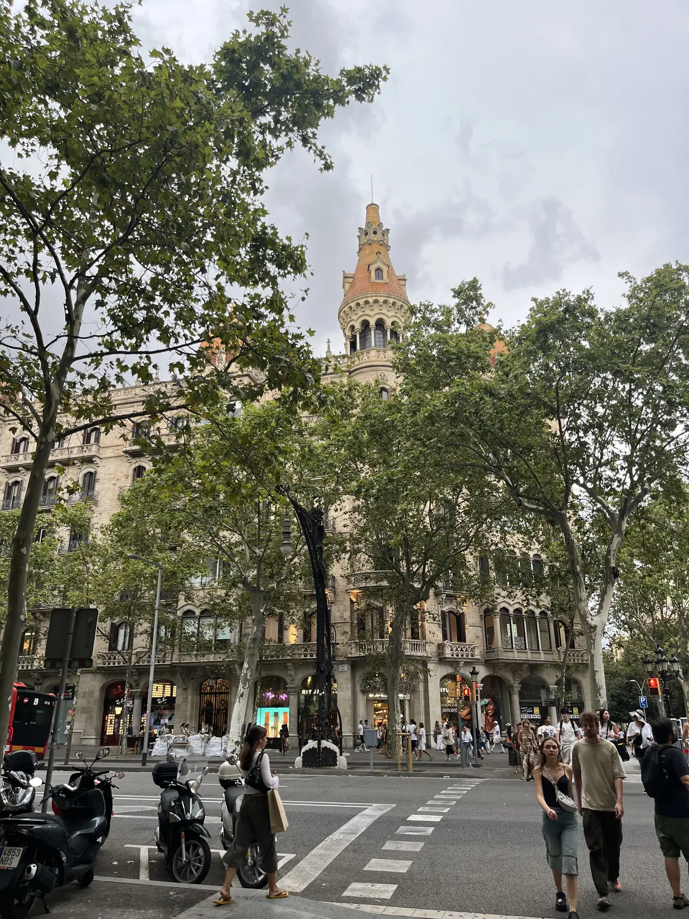An ornate modernist tower with a conical roof rising above a Passeig de Gràcia building in Barcelona, trees framing the view