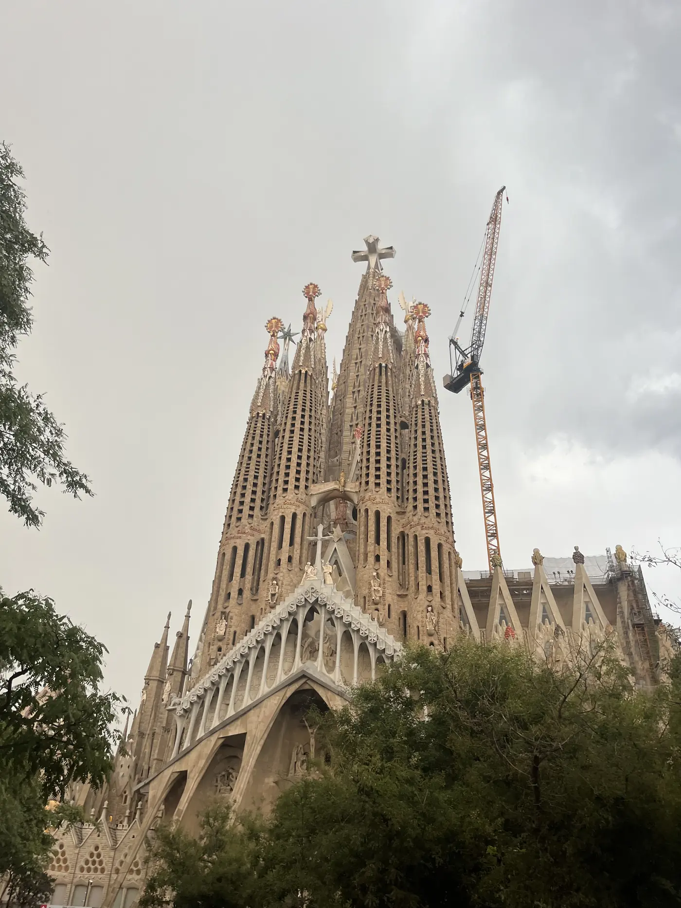 The towering spires of the Sagrada Família under an overcast sky, construction crane visible beside the unfinished basilica