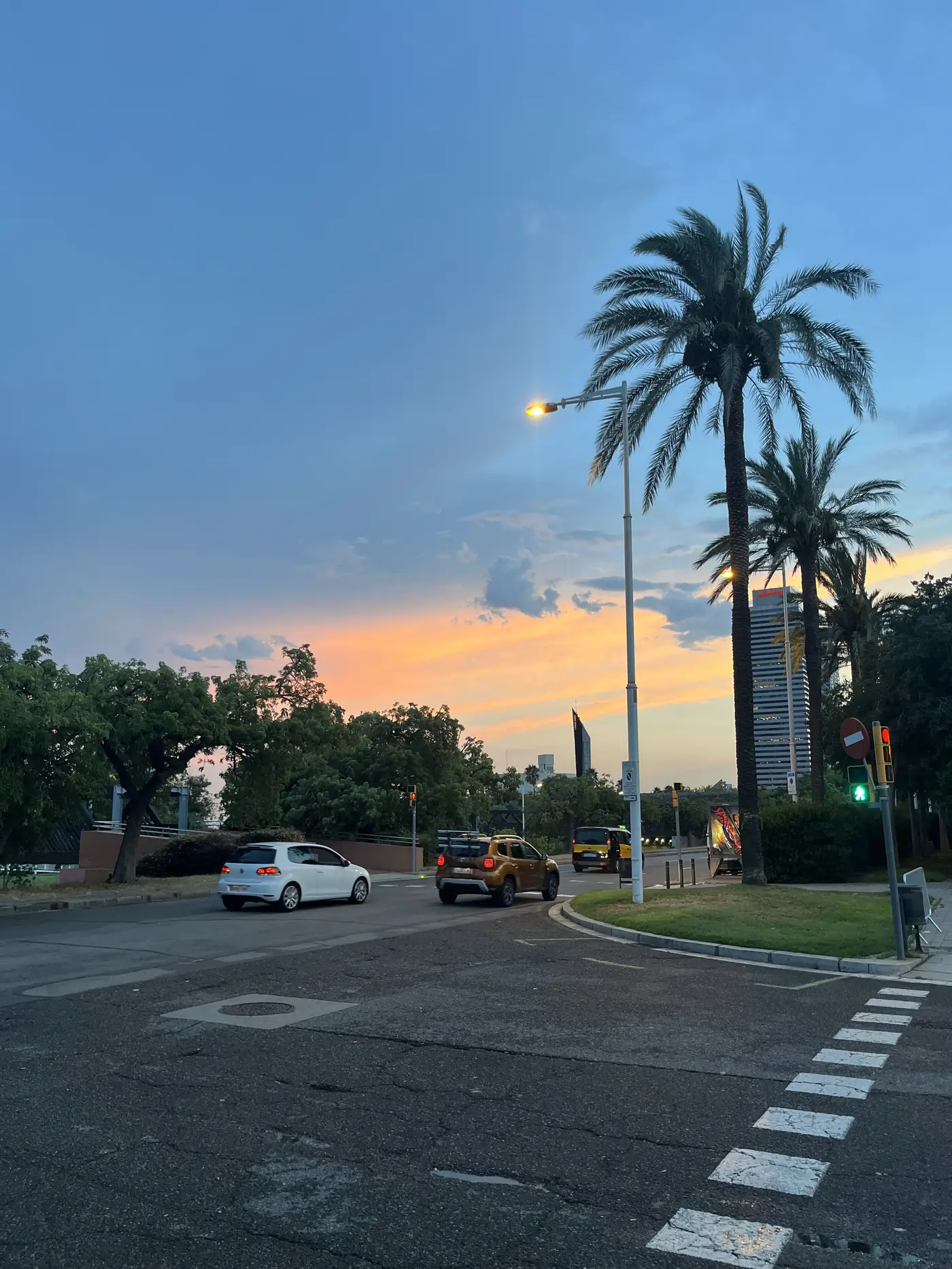 Palm trees silhouetted against a golden Barcelona sunset over a quiet intersection, cars waiting at a crossing