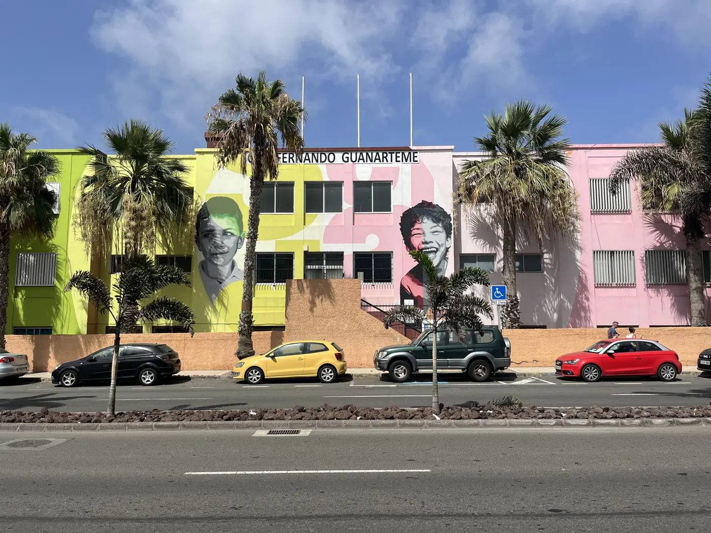 Colourful street mural of faces on a building facade in the Fernando Guanarteme neighbourhood of Las Palmas, palm trees and parked cars in front