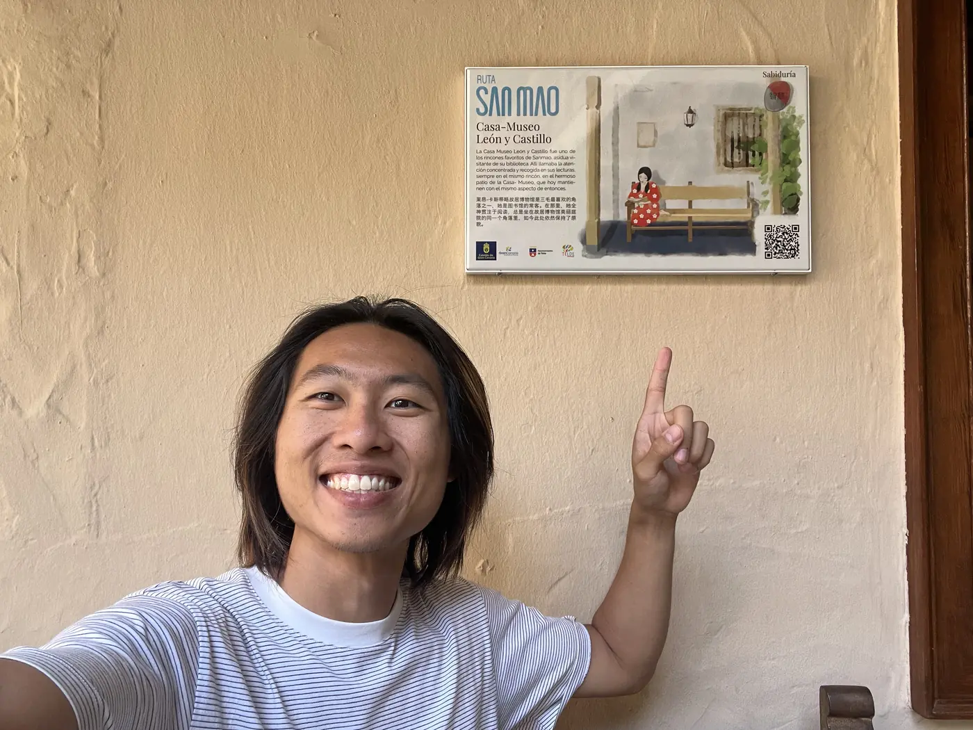 Shin pointing up at a Sanmao commemorative plaque mounted on a stucco wall in a courtyard in Gran Canaria