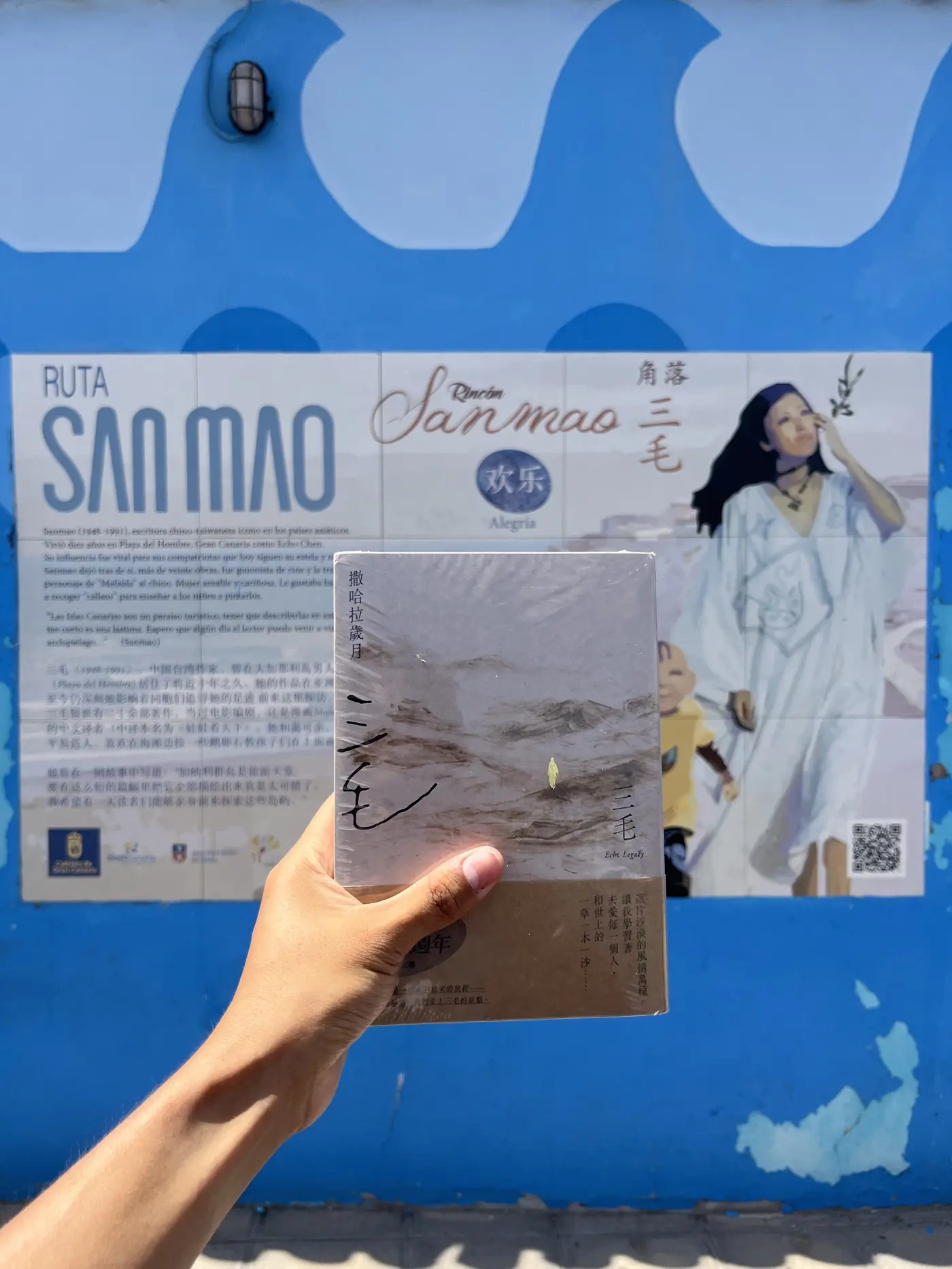 A hand holding a copy of Sanmao's book Stories of the Sahara in front of a blue mural illustrating her life, Gran Canaria