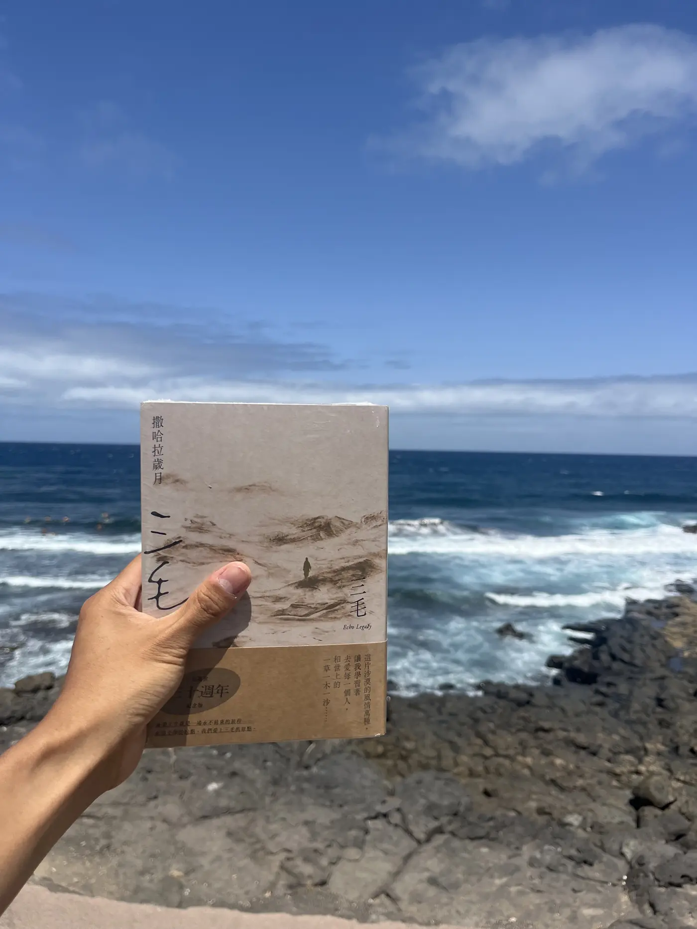 A hand holding a copy of Sanmao's book Stories of the Sahara against a backdrop of crashing Atlantic waves and volcanic rock in Gran Canaria