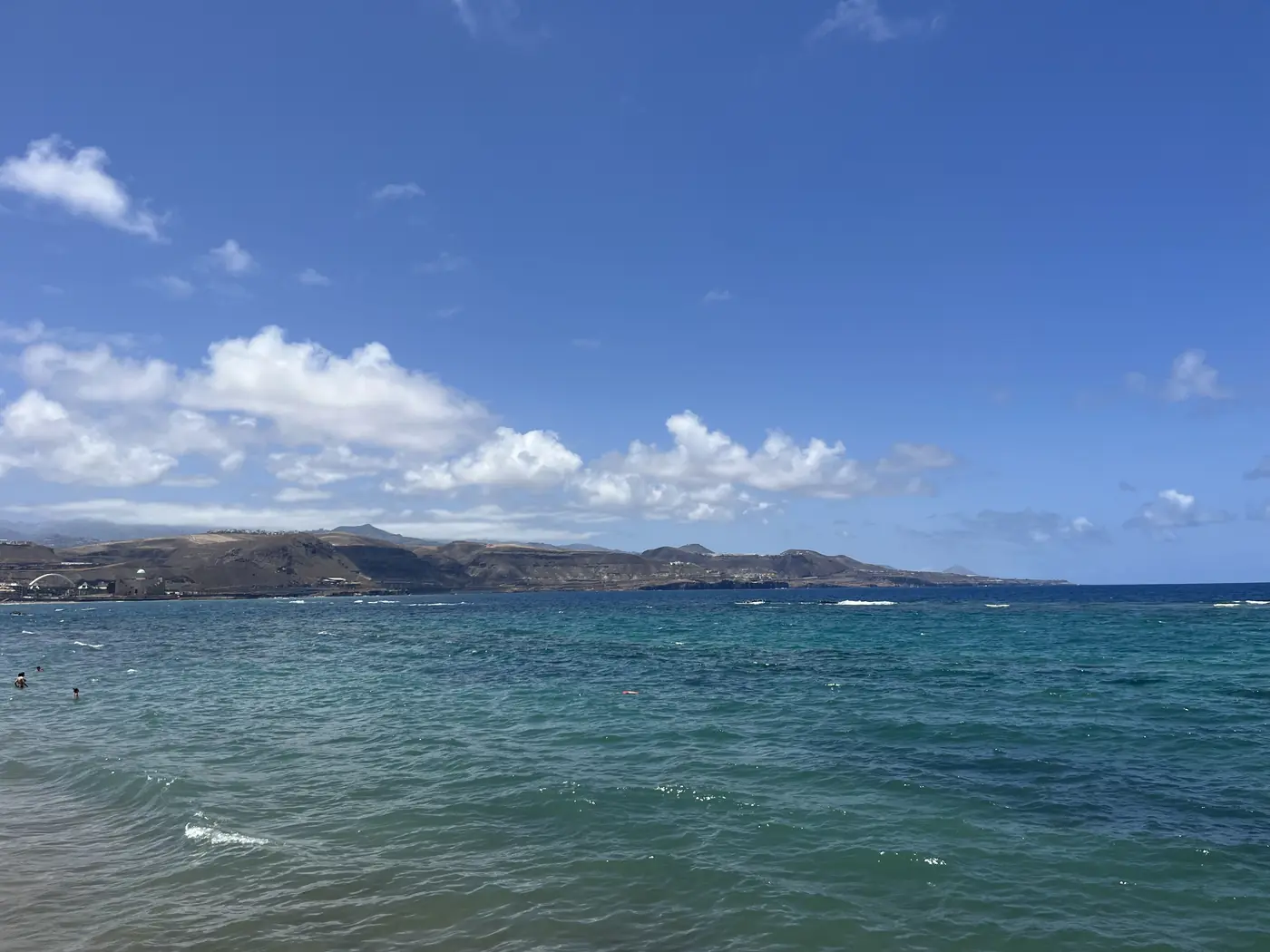 Calm turquoise ocean water with the Gran Canaria coastline and hills visible in the distance under a clear sky