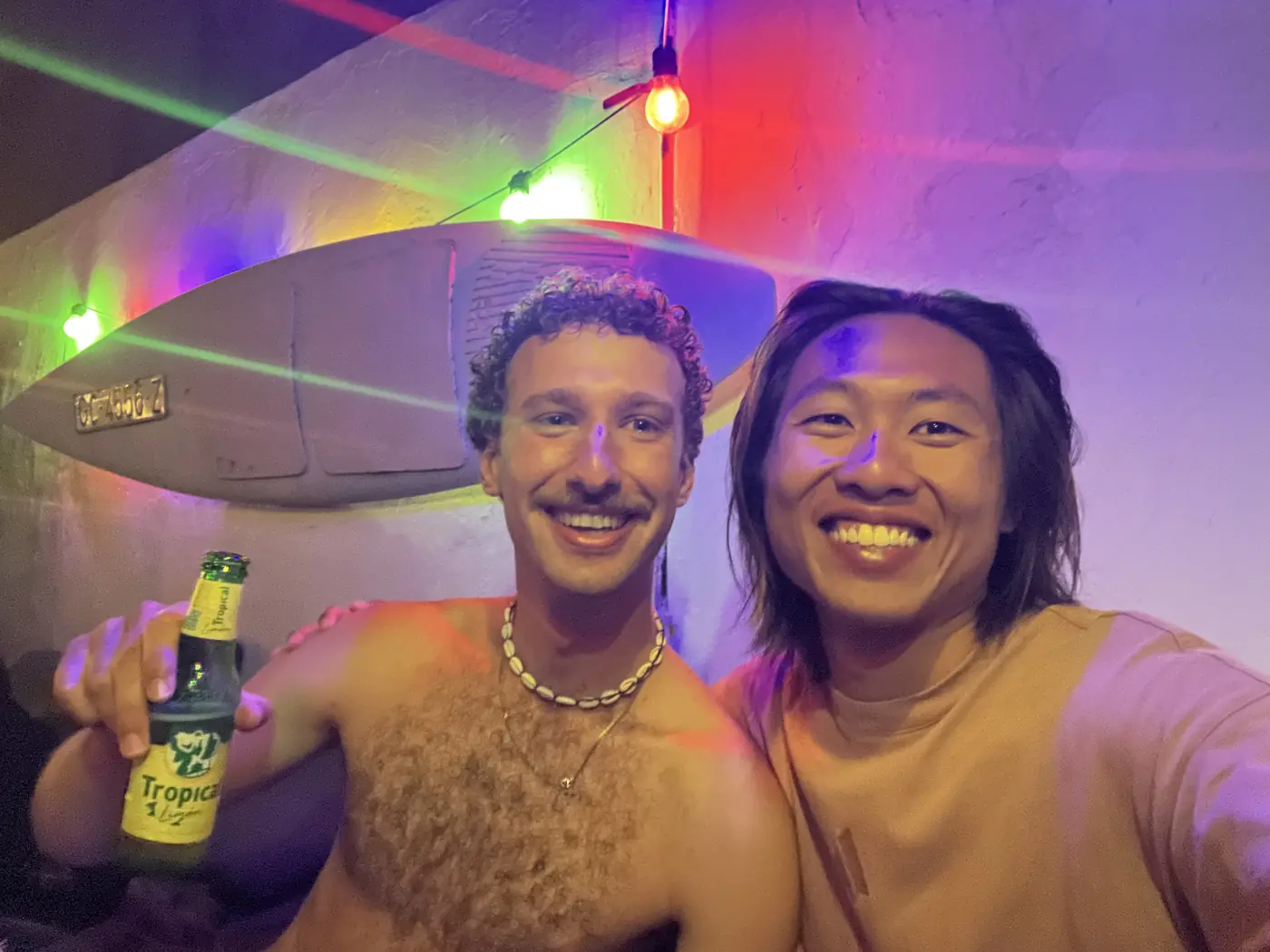 Shin and a friend smiling at a colourful nighttime rooftop party in Gran Canaria, string lights and a beer bottle in hand