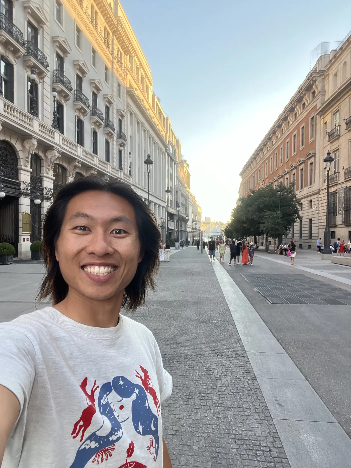 Shin selfie on a wide pedestrian boulevard in central Madrid at golden hour, grand stone buildings lining both sides