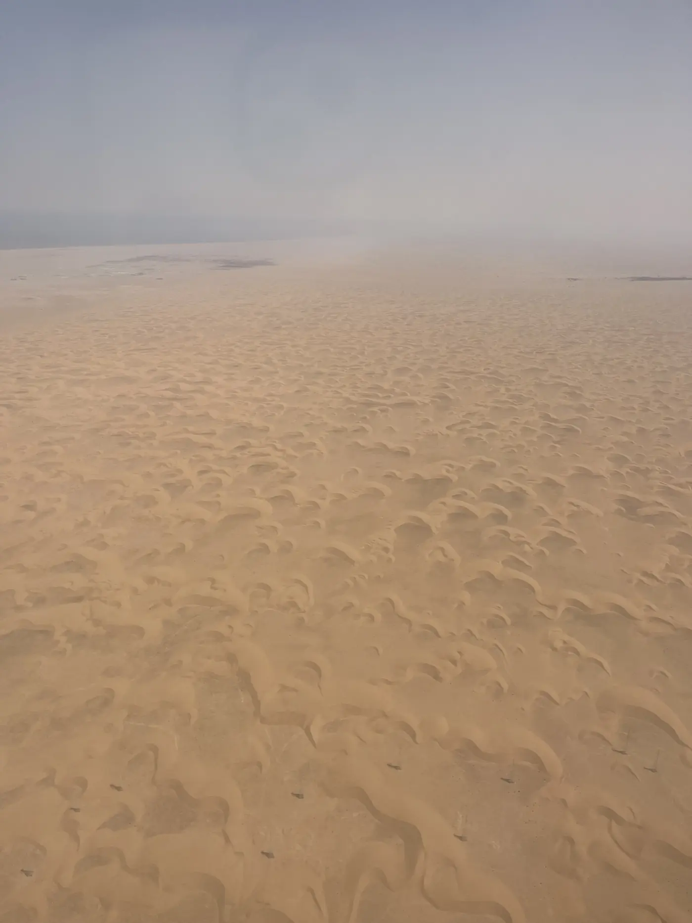 Vast hazy Sahara sand dunes stretching to the horizon, seen from above through a plane window