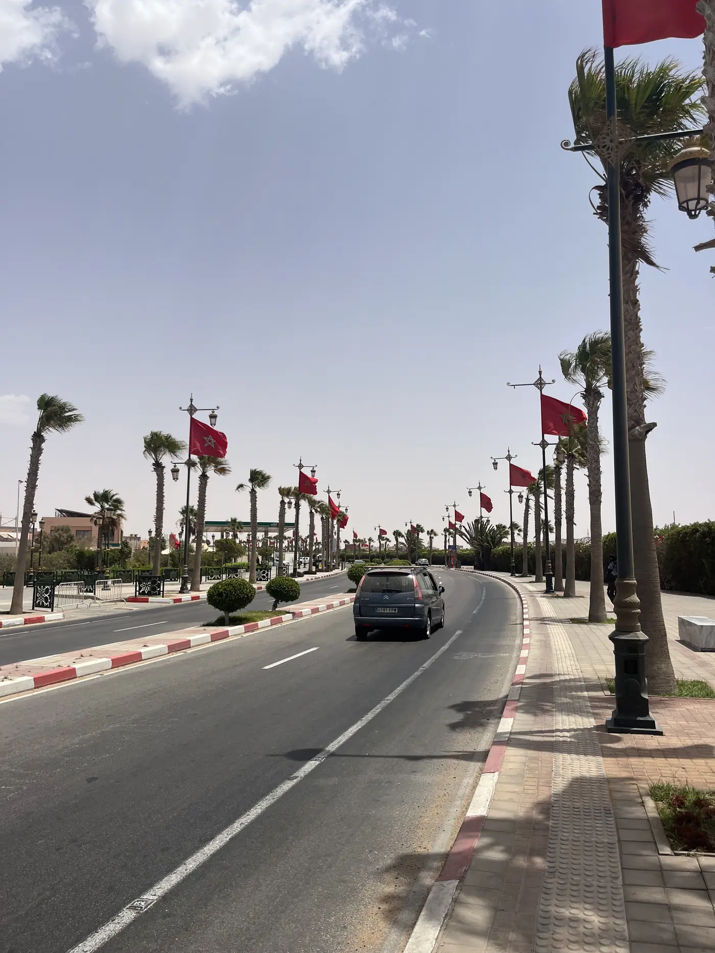 A palm tree-lined boulevard in Laayoune decorated with Moroccan flags on lampposts, a car driving down the empty road