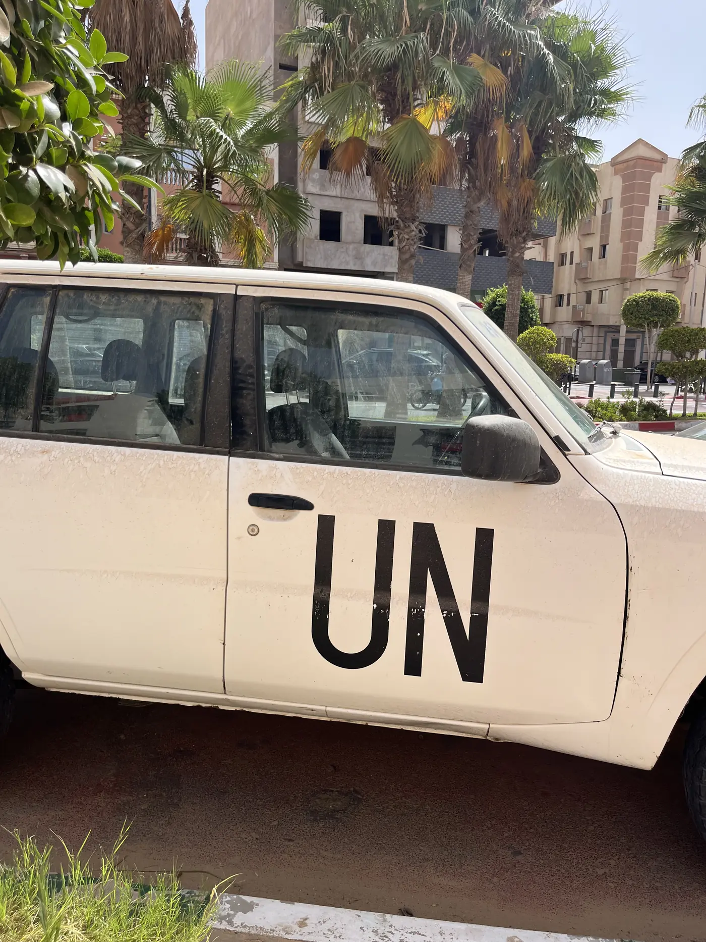 A white UN-marked vehicle parked on a street in Laayoune, palm trees and buildings behind it
