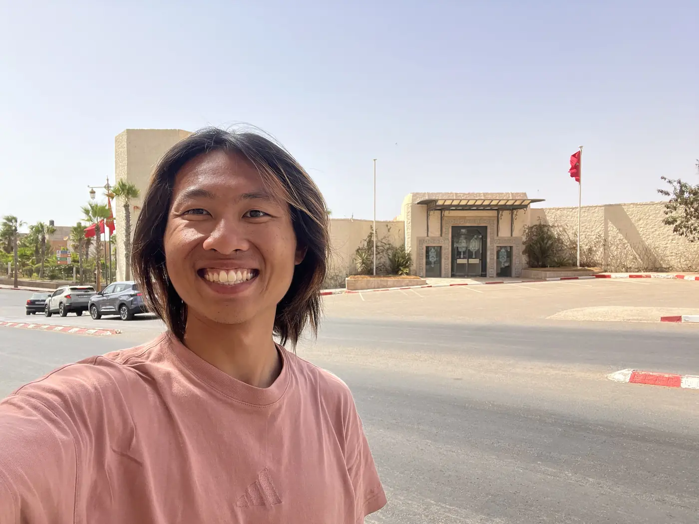 Shin selfie in front of a modern sand-coloured building with a Moroccan flag, palm trees along the street in Laayoune