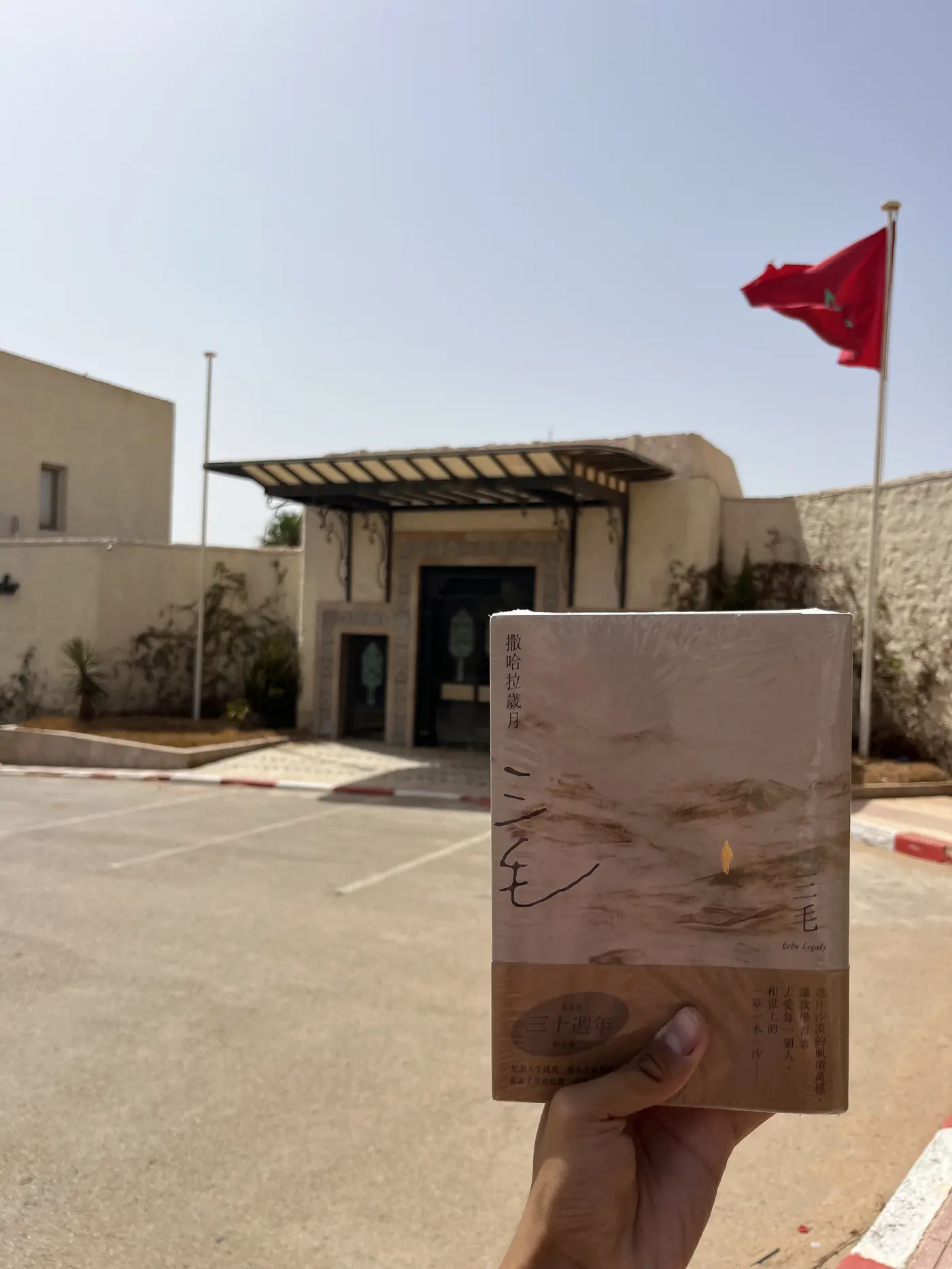 A hand holding a copy of Sanmao's book Stories of the Sahara in front of a building with a Moroccan flag in Laayoune
