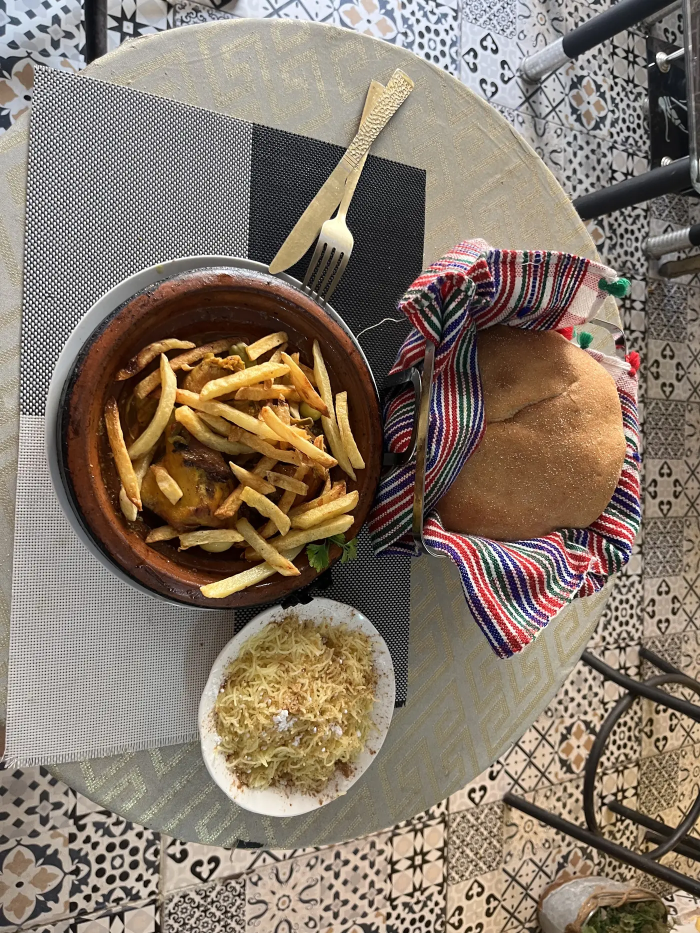Overhead view of a Sahrawi meal in Laayoune — a clay tagine of fries and meat, flatbread wrapped in cloth, and a plate of seffa