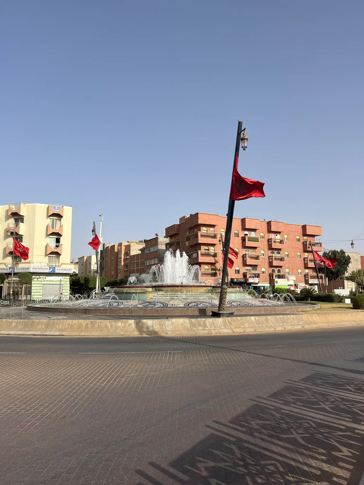 A fountain roundabout in central Laayoune with red Moroccan flags and apartment buildings around the plaza