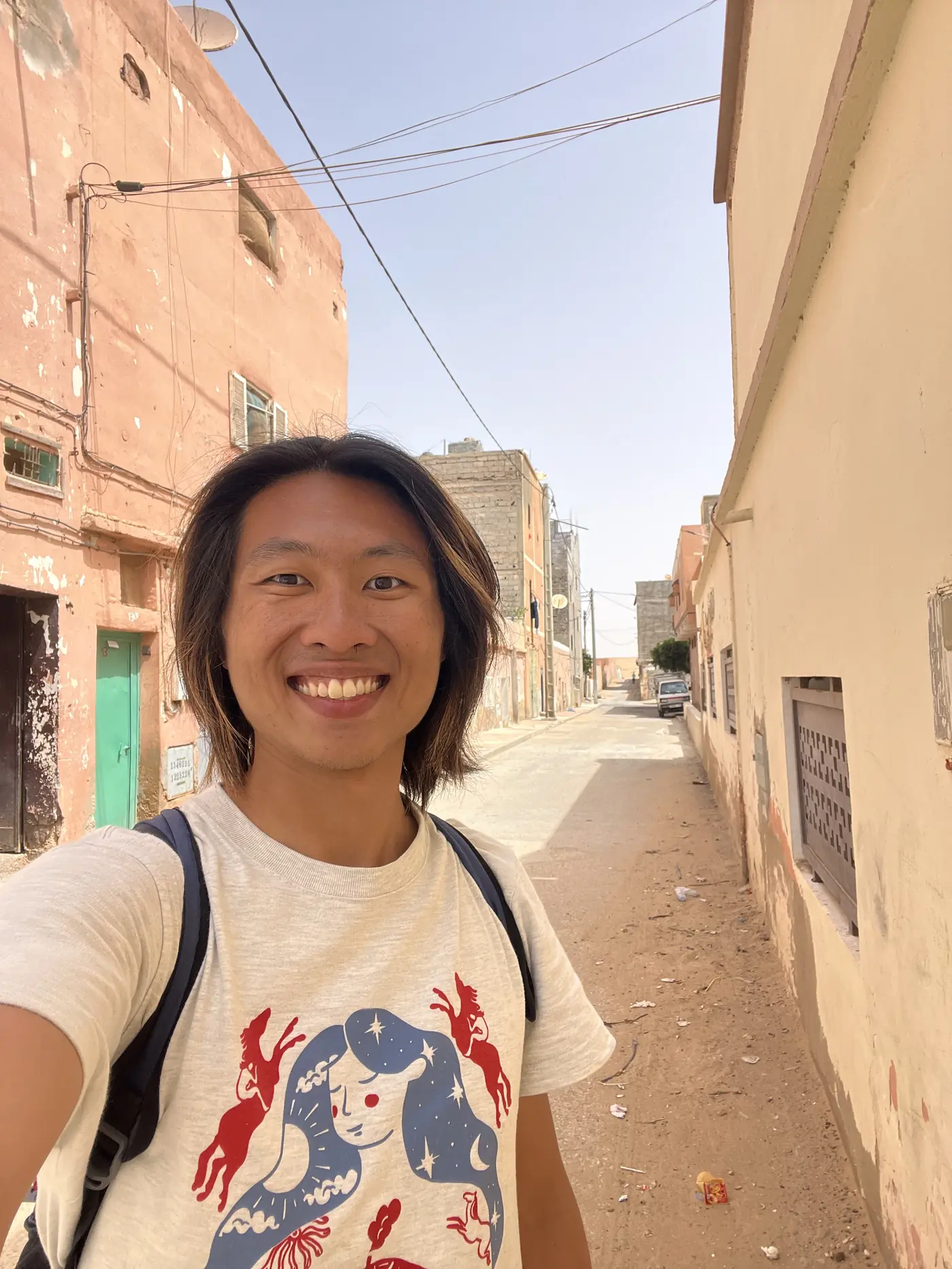 Shin smiling in the same narrow old-town alley in Laayoune, weathered buildings on either side