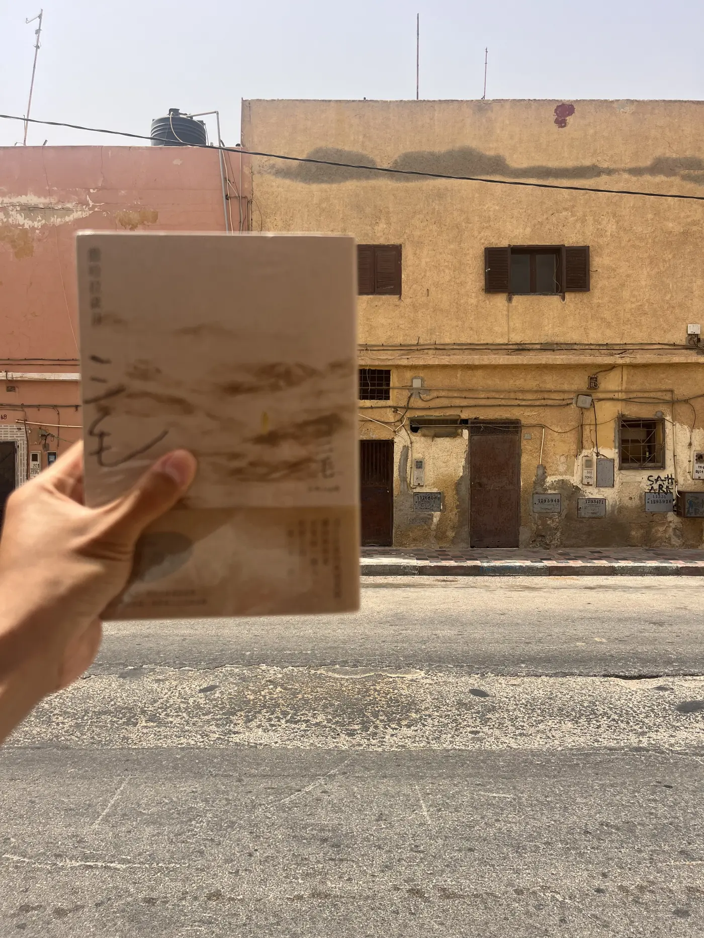 A hand holding a copy of Sanmao's book Stories of the Sahara in front of a weathered Spanish colonial-era building in Laayoune
