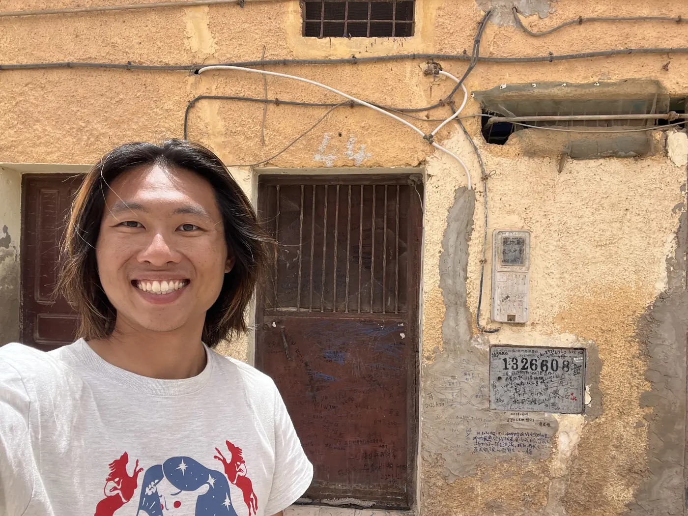 Shin smiling in front of an old weathered door with a faded address plaque in Laayoune's old town