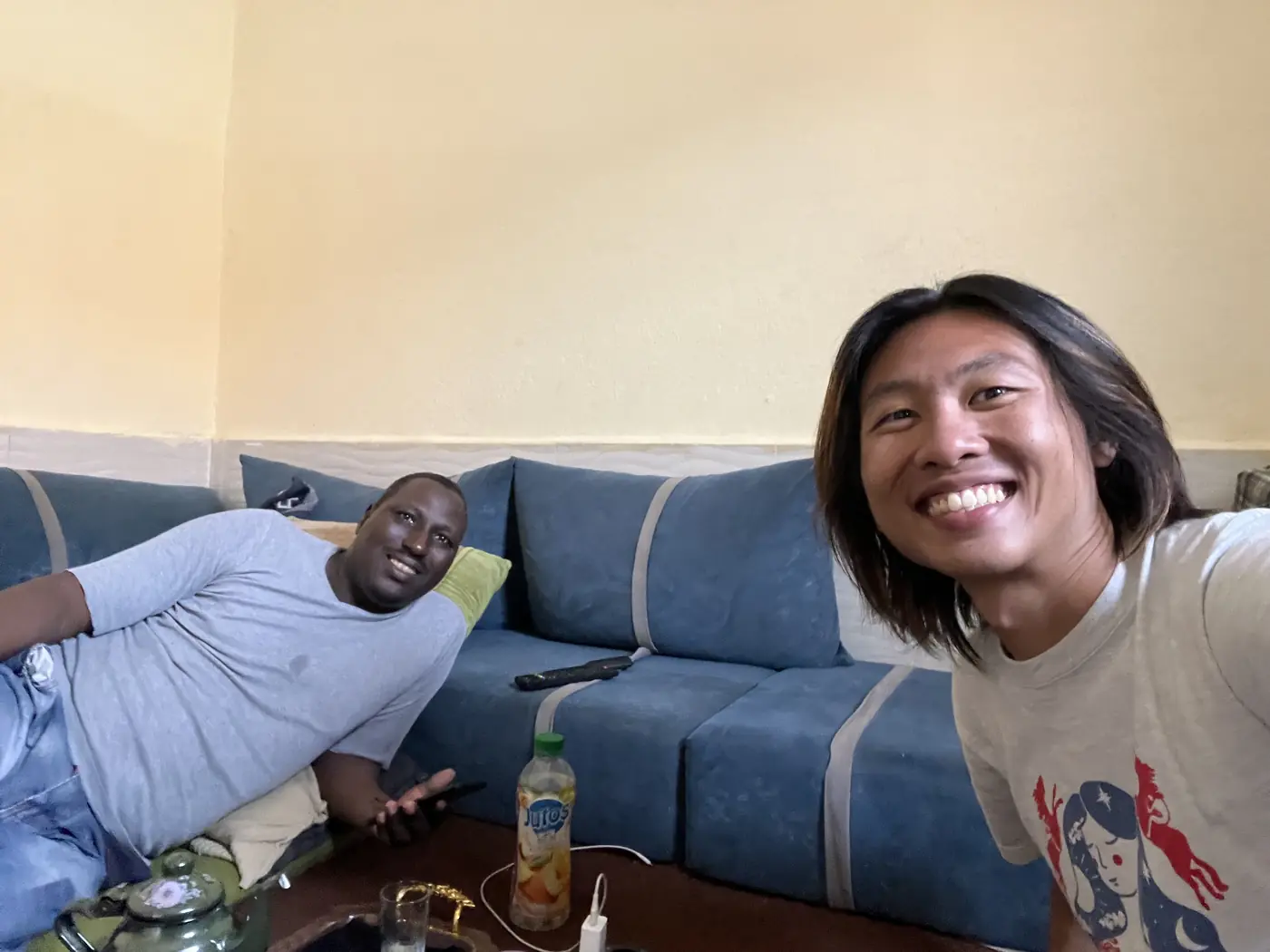 Shin and a local friend sitting together on a blue sofa in a Laayoune living room, drinks on the table