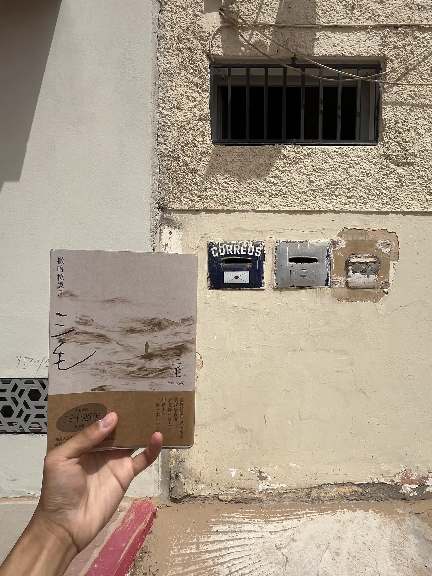 A hand holding a copy of Sanmao's book Stories of the Sahara beside an old Spanish 'Correos' postal box mounted on a wall in Laayoune